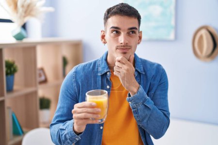 Foto de Hombre hispano guapo bebiendo vaso de jugo de naranja cara seria pensando en la pregunta con la mano en la barbilla, pensativo acerca de la idea confusa - Imagen libre de derechos