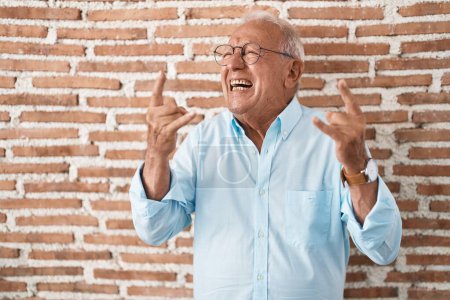 Téléchargez les photos : Senior man with grey hair standing over bricks wall shouting with crazy expression doing rock symbol with hands up. music star. heavy music concept. - en image libre de droit