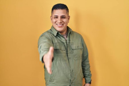 Foto de Hispanic young man standing over yellow background smiling friendly offering handshake as greeting and welcoming. successful business. - Imagen libre de derechos