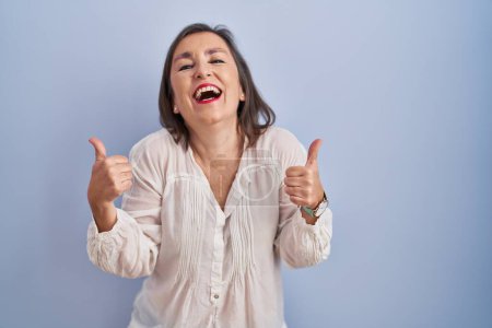 Foto de Mujer hispana de mediana edad de pie sobre el signo de éxito de fondo azul haciendo gesto positivo con la mano, pulgares hacia arriba sonriendo y feliz. expresión alegre y gesto ganador. - Imagen libre de derechos
