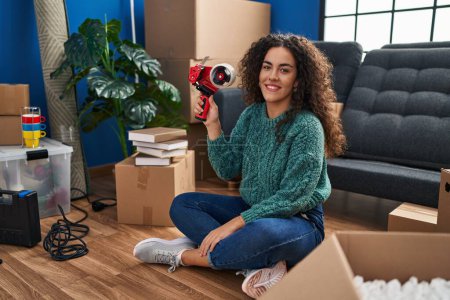 Foto de Young beautiful hispanic woman smiling confident holding package tape machine at new home - Imagen libre de derechos