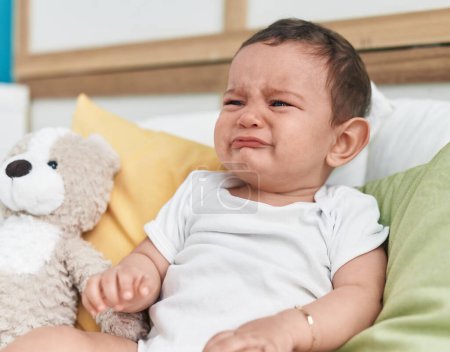 Foto de Adorable bebé hispano sentado en la cama llorando en el dormitorio - Imagen libre de derechos