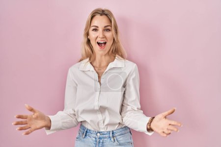 Foto de Mujer caucásica joven con camisa blanca casual sobre fondo rosa sonriente alegre con los brazos abiertos como bienvenida amistosa, saludos positivos y confiados - Imagen libre de derechos