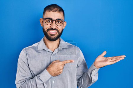 Téléchargez les photos : Middle east man with beard standing over blue background amazed and smiling to the camera while presenting with hand and pointing with finger. - en image libre de droit