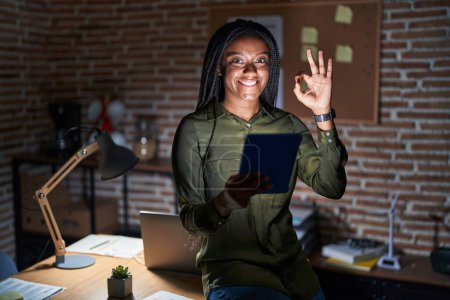 Photo for Young african american with braids working at the office at night smiling positive doing ok sign with hand and fingers. successful expression. - Royalty Free Image