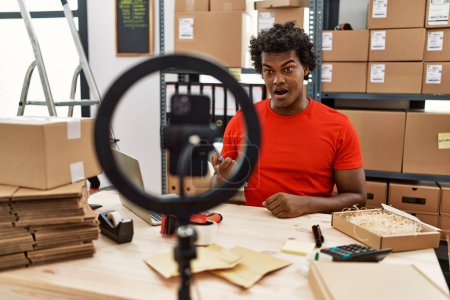 Foto de African man working at small business ecommerce doing video tutorial scared and amazed with open mouth for surprise, disbelief face - Imagen libre de derechos