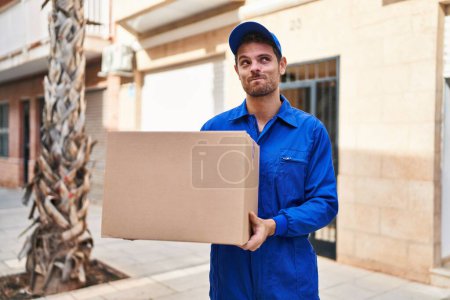 Foto de Young hispanic man delivering box smiling looking to the side and staring away thinking. - Imagen libre de derechos
