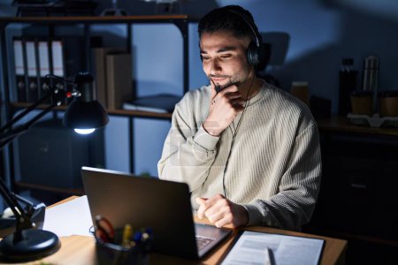 Photo for Young handsome man working using computer laptop at night looking confident at the camera with smile with crossed arms and hand raised on chin. thinking positive. - Royalty Free Image
