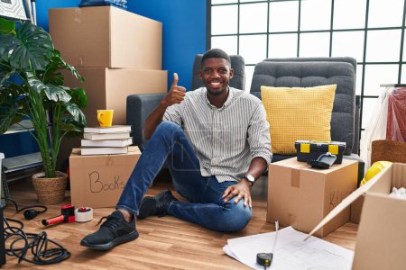 Foto de African american man sitting on the floor at new home doing happy thumbs up gesture with hand. approving expression looking at the camera showing success. - Imagen libre de derechos