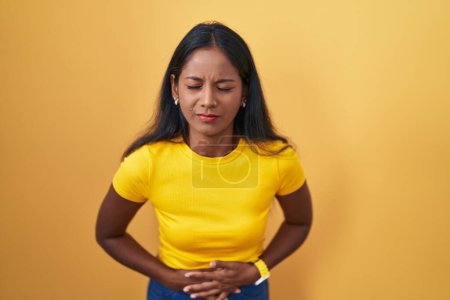 Téléchargez les photos : Young indian woman standing over yellow background with hand on stomach because indigestion, painful illness feeling unwell. ache concept. - en image libre de droit