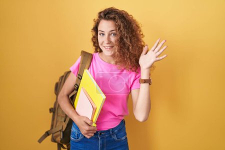Foto de Young caucasian woman wearing student backpack and holding books waiving saying hello happy and smiling, friendly welcome gesture - Imagen libre de derechos