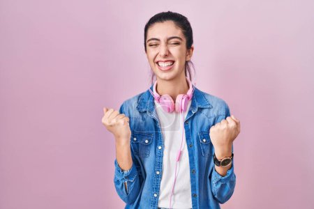 Foto de Young beautiful woman standing over pink background very happy and excited doing winner gesture with arms raised, smiling and screaming for success. celebration concept. - Imagen libre de derechos