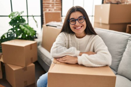 Téléchargez les photos : Jeune femme hispanique souriant confiant appuyé sur le paquet à la nouvelle maison - en image libre de droit