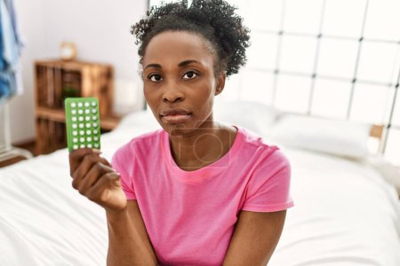 Foto de Mujer afroamericana sosteniendo píldoras anticonceptivas sentada en la cama en el dormitorio - Imagen libre de derechos