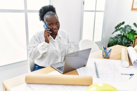 Photo for Young african american woman architect using laptop and talking on the smartphone at architecture studio - Royalty Free Image