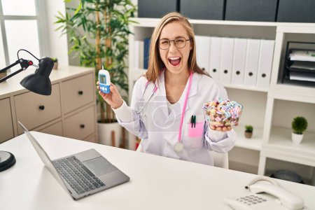 Téléchargez les photos : Young blonde doctor woman holding glucometer and sweets smiling and laughing hard out loud because funny crazy joke. - en image libre de droit