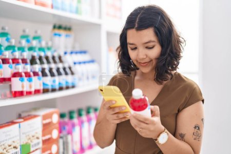 Téléchargez les photos : Jeune femme client utilisant smartphone tenant bouteille de médicaments à la pharmacie - en image libre de droit
