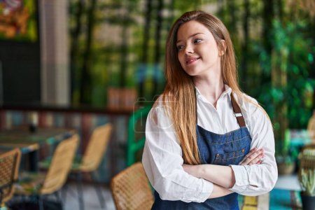 Foto de Joven mujer caucásica camarera sonriendo confiada de pie con los brazos cruzados gesto en el restaurante - Imagen libre de derechos