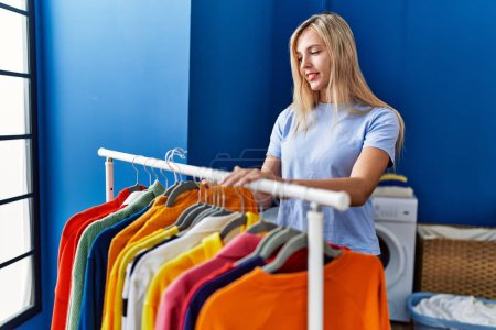 Photo for Young blonde woman smiling confident hanging clothes on rack at laundry room - Royalty Free Image