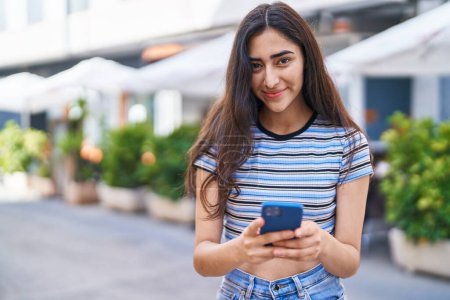 Téléchargez les photos : Jeune fille hispanique souriant confiant en utilisant smartphone à la rue - en image libre de droit