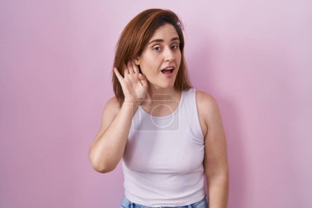Photo for Brunette woman standing over pink background smiling with hand over ear listening an hearing to rumor or gossip. deafness concept. - Royalty Free Image
