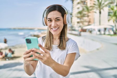 Téléchargez les photos : Jeune femme hispanique souriant confiant en utilisant smartphone et écouteurs au bord de la mer - en image libre de droit