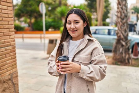 Téléchargez les photos : Jeune femme hispanique souriant boire du café en toute confiance dans la rue - en image libre de droit