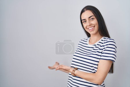 Foto de Young brunette woman wearing striped t shirt inviting to enter smiling natural with open hand - Imagen libre de derechos