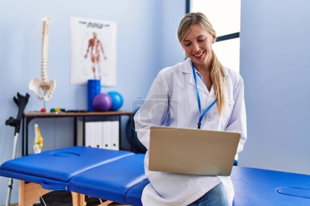 Téléchargez les photos : Jeune femme blonde portant un uniforme de physiothérapeute utilisant un ordinateur portable à la clinique de physiothérapie - en image libre de droit