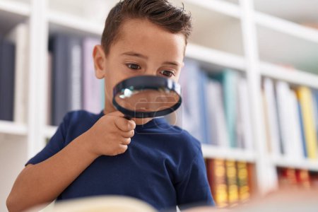 Photo for Adorable hispanic toddler student reading book using magnifying glass at library school - Royalty Free Image