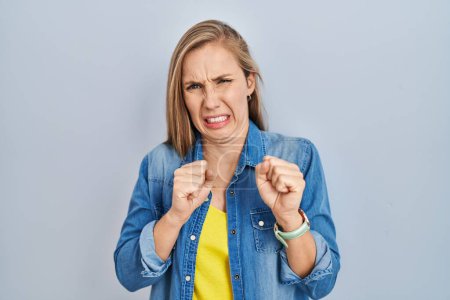 Foto de Young blonde woman standing over blue background disgusted expression, displeased and fearful doing disgust face because aversion reaction. - Imagen libre de derechos