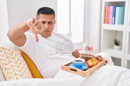 Téléchargez les photos : Jeune homme hispanique mangeant le petit déjeuner dans le lit avec le visage en colère, signe négatif montrant aversion avec les pouces vers le bas, concept de rejet - en image libre de droit