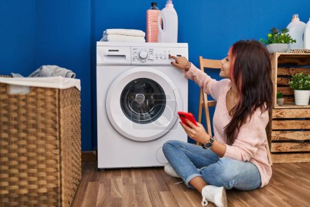 Téléchargez les photos : Jeune femme caucasienne utilisant smartphone allumant la machine à laver à la buanderie - en image libre de droit