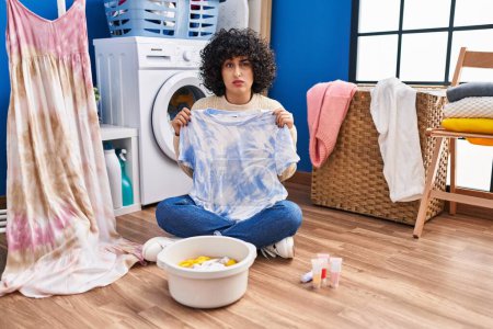 Foto de Young brunette woman with curly hair dyeing tye die t shirt and dress skeptic and nervous, frowning upset because of problem. negative person. - Imagen libre de derechos