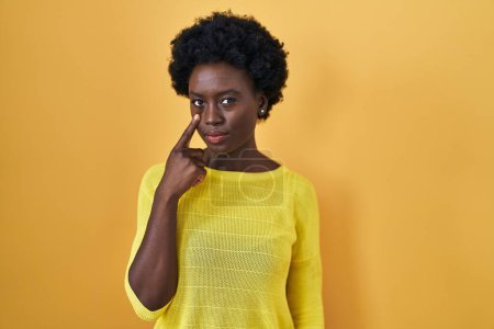 Téléchargez les photos : Jeune femme africaine debout au-dessus de studio jaune pointant vers l'oeil vous regarder geste, expression suspecte - en image libre de droit