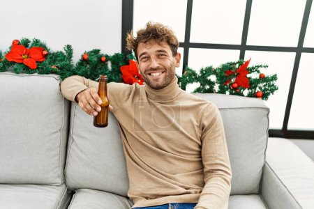 Téléchargez les photos : Jeune homme hispanique buvant de la bière assis près du décor de Noël à la maison - en image libre de droit