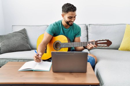 Téléchargez les photos : Jeune homme arabe composer chanson à l'aide d'un ordinateur portable et guitare classique à la maison - en image libre de droit