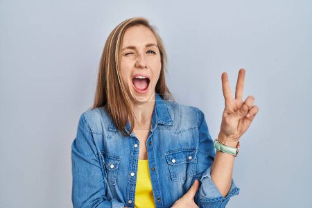 Téléchargez les photos : Young blonde woman standing over blue background smiling with happy face winking at the camera doing victory sign with fingers. number two. - en image libre de droit