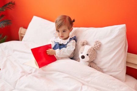 Téléchargez les photos : Adorable livre de lecture blond pour tout-petit assis sur le lit avec un ours en peluche dans la chambre - en image libre de droit