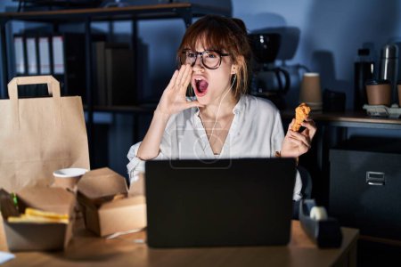Photo for Young beautiful woman working using computer laptop and eating delivery food shouting and screaming loud to side with hand on mouth. communication concept. - Royalty Free Image