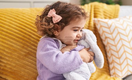 Foto de Adorable chica hispana abrazando conejo juguete sentado en sofá en casa - Imagen libre de derechos