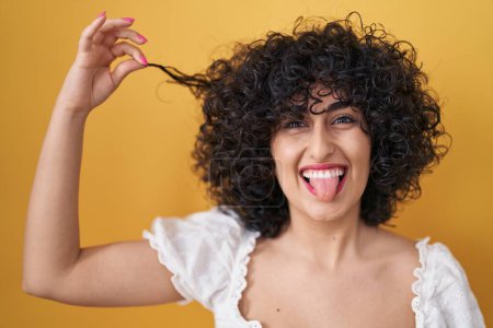 Foto de Young brunette woman with curly hair holding curl sticking tongue out happy with funny expression. - Imagen libre de derechos