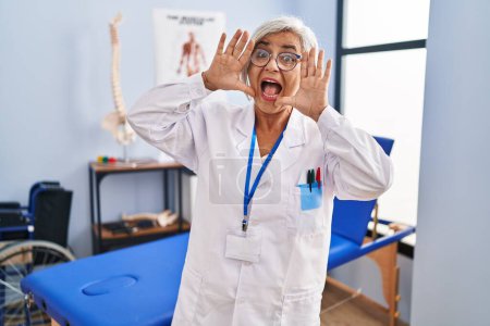 Foto de Middle age woman with grey hair working at pain recovery clinic smiling cheerful playing peek a boo with hands showing face. surprised and exited - Imagen libre de derechos