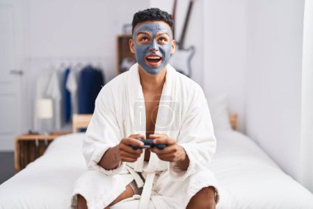 Foto de Young hispanic man wearing beauty face mask playing video games angry and mad screaming frustrated and furious, shouting with anger looking up. - Imagen libre de derechos