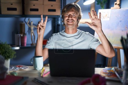 Téléchargez les photos : Middle age man sitting at art studio with laptop at night showing and pointing up with fingers number seven while smiling confident and happy. - en image libre de droit