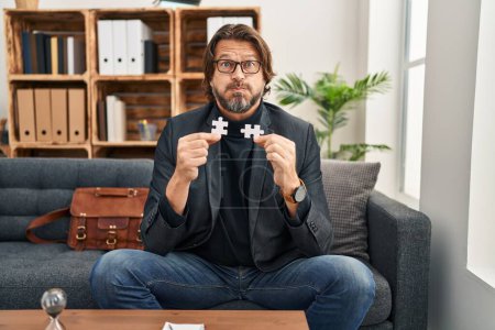 Photo for Handsome middle age man at consultation office holding puzzle pieces puffing cheeks with funny face. mouth inflated with air, catching air. - Royalty Free Image