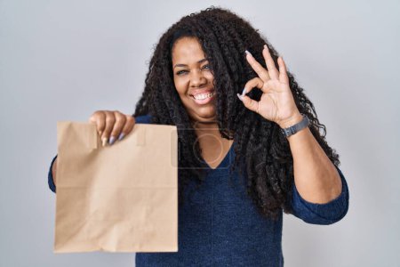 Foto de Plus size hispanic woman holding take away paper bag doing ok sign with fingers, smiling friendly gesturing excellent symbol - Imagen libre de derechos