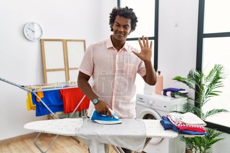 Téléchargez les photos : African man with curly hair ironing clothes at home waiving saying hello happy and smiling, friendly welcome gesture - en image libre de droit