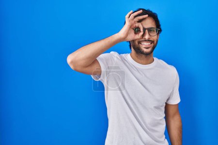 Photo for Handsome latin man standing over blue background doing ok gesture with hand smiling, eye looking through fingers with happy face. - Royalty Free Image