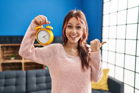 Photo for Young caucasian woman holding alarm clock pointing thumb up to the side smiling happy with open mouth - Royalty Free Image